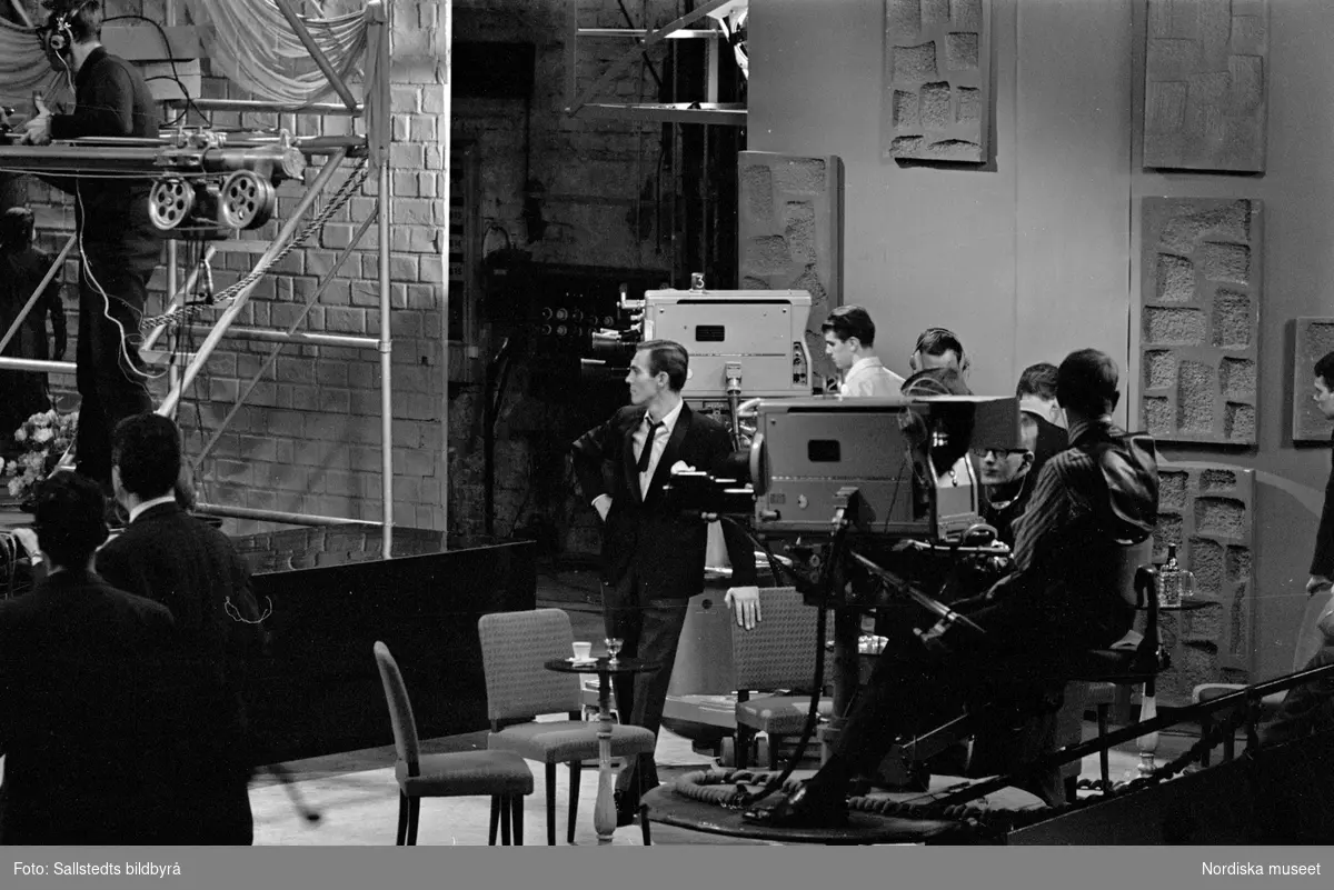 Foto från TV-inspelning på Cirkus med Gunnar Wiklund. Troligen inspelning av "Gunnar Wiklund show". Gunnar Wiklund står vid en stol omgiven av tv-kameror.  Statister och inspelningspersonal står runtomkring i studion.