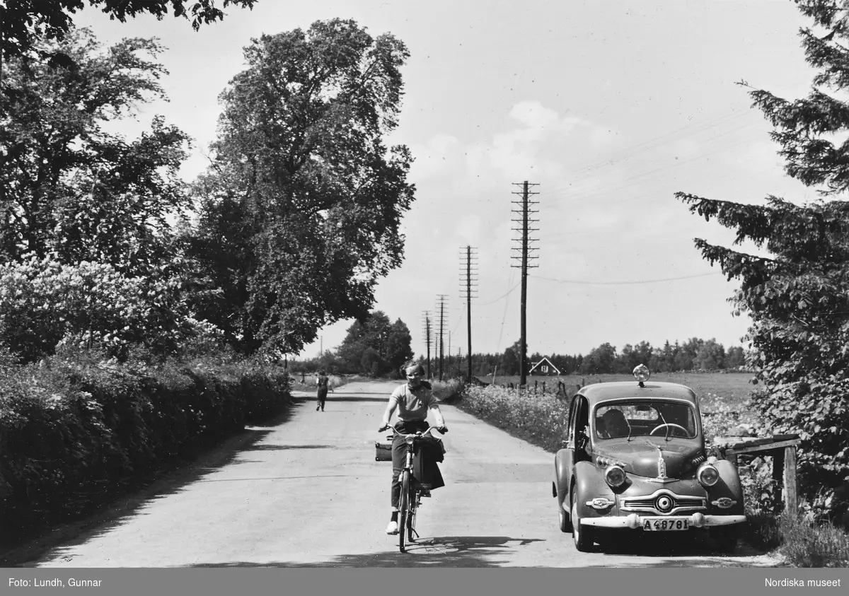 Landsvägsvy från Skåne, 1955. En flicka cyklar förbi fotograf Gunnar Lundhs parkerade bil, en Dyna Panhard.