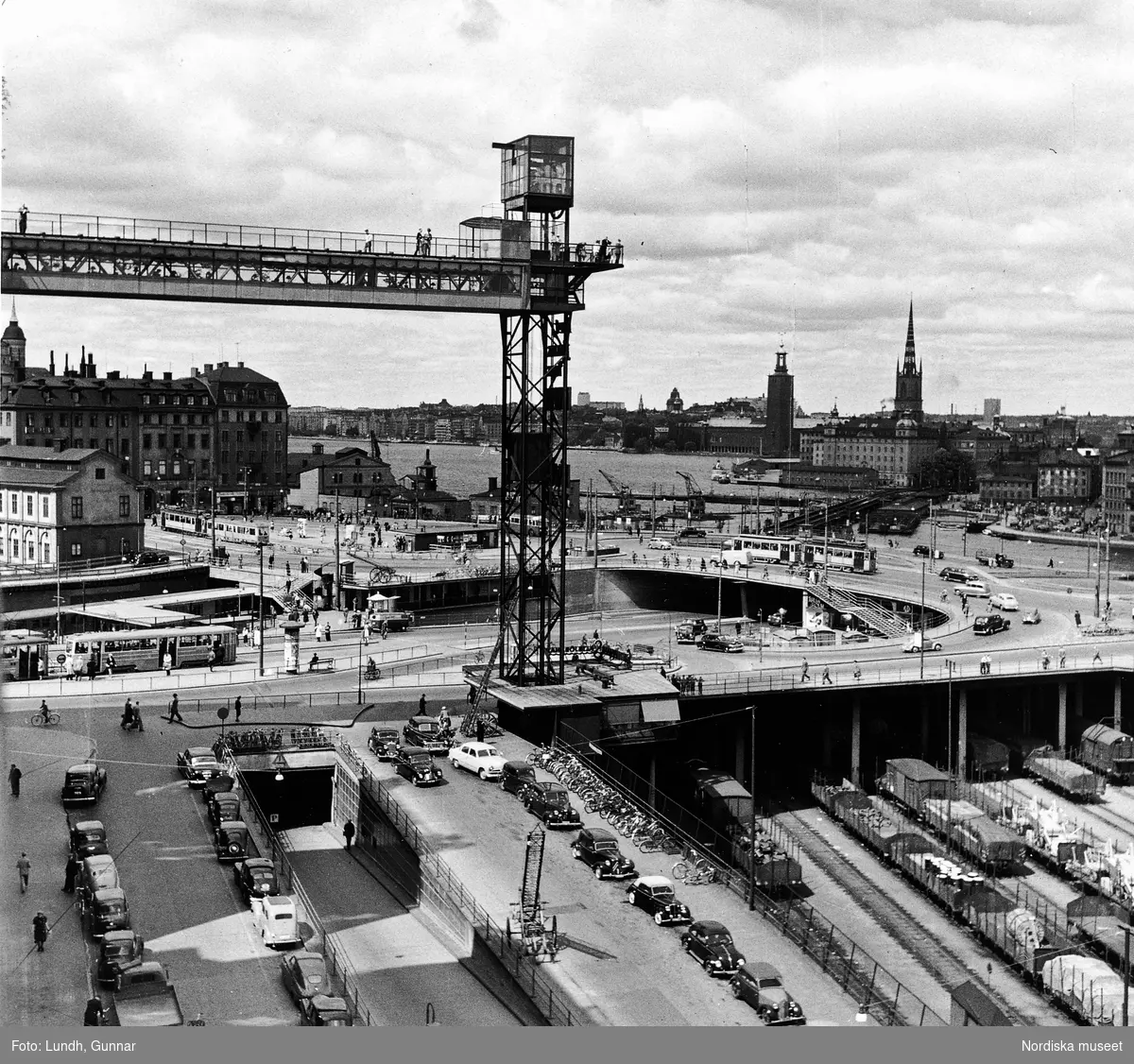 Vy över Slussen och Katarinahissen, Stockholm, år 1951. Parkerade bilar, cyklar, järnvägsvagnar i förgrunden.