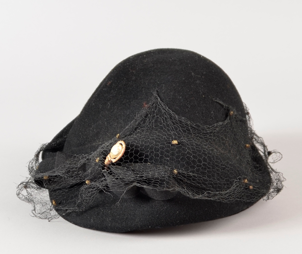 Hatt. Svart filt. Mjuk. Framme svart nett med "gullklumpar" over svart filtsløyfe med hattenål med stor imitert perle i gyllen innfatting. Strikk under til å ha under haka. Ufôra.