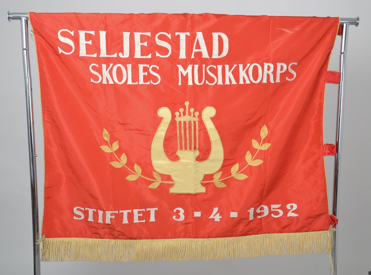 Fane fra Seljestad Skoles Musikkorps i Harstad i rød silke med gullsnorer, applisert og brodert med hvit og gul tråd.