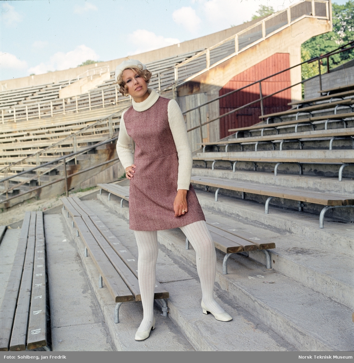 Motebilde, kvinne i lilla ullkjole med hvit genser, sko, strømper og alpelue, på tribunen på idrettsarena. Bildet er en del av en serie der noen er publisert i ukebladet Det Nye 1969, nr. 38.