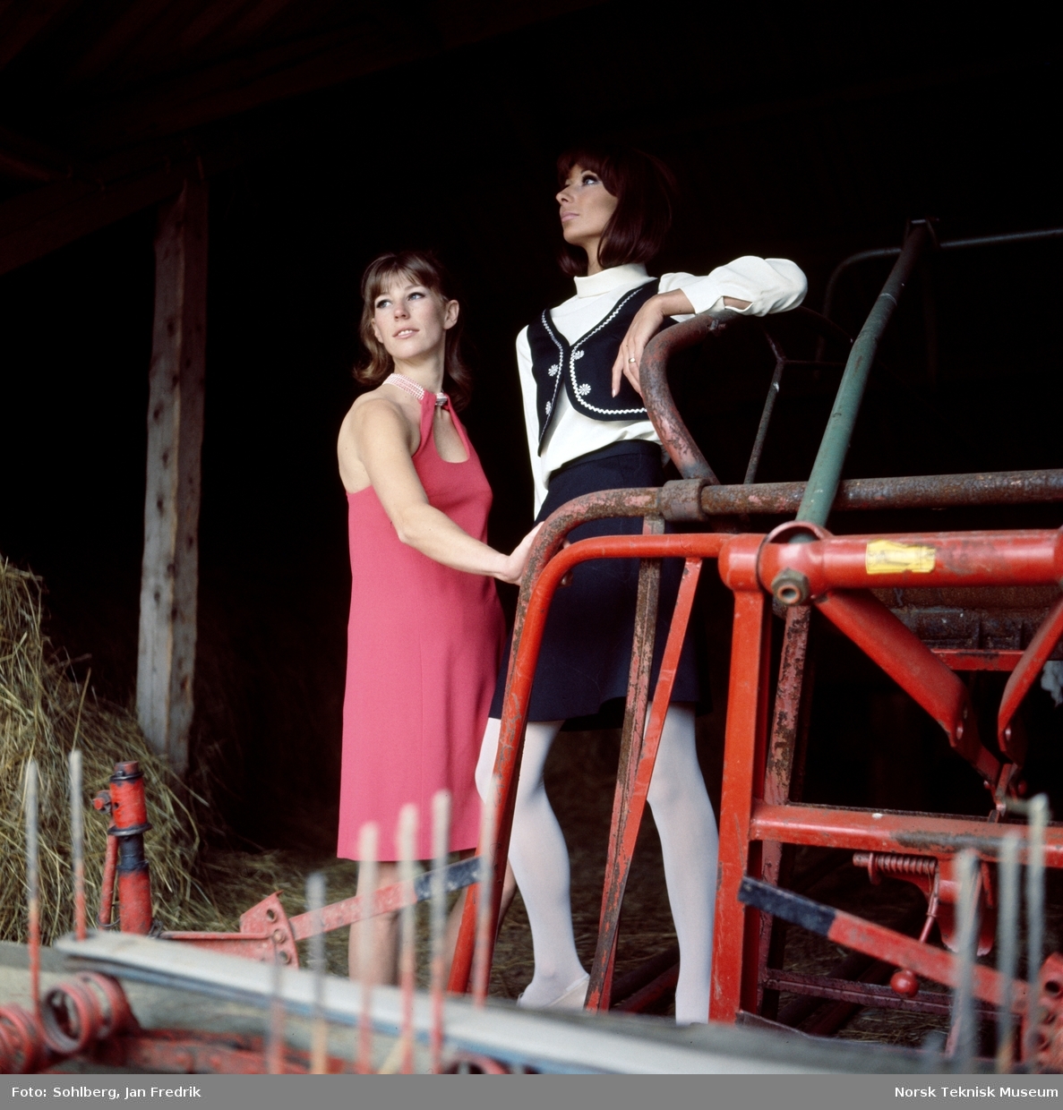 To kvinnelige modeller, den ene i rød kjole og den andre i sort vest og skjørt med hvit bluse inni fotografert på låven ved landbruksredskaper, høsten 1968.