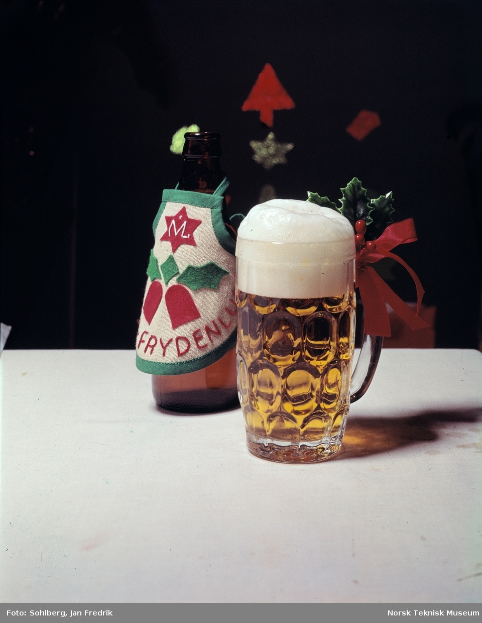 Humoristisk reklame for juleøl av merket Frydenlund. En ølflaske har fått et forkle med julemotiv og Frydenlunds logo rundt seg. Foran flaska står en seidel med skummende øl.