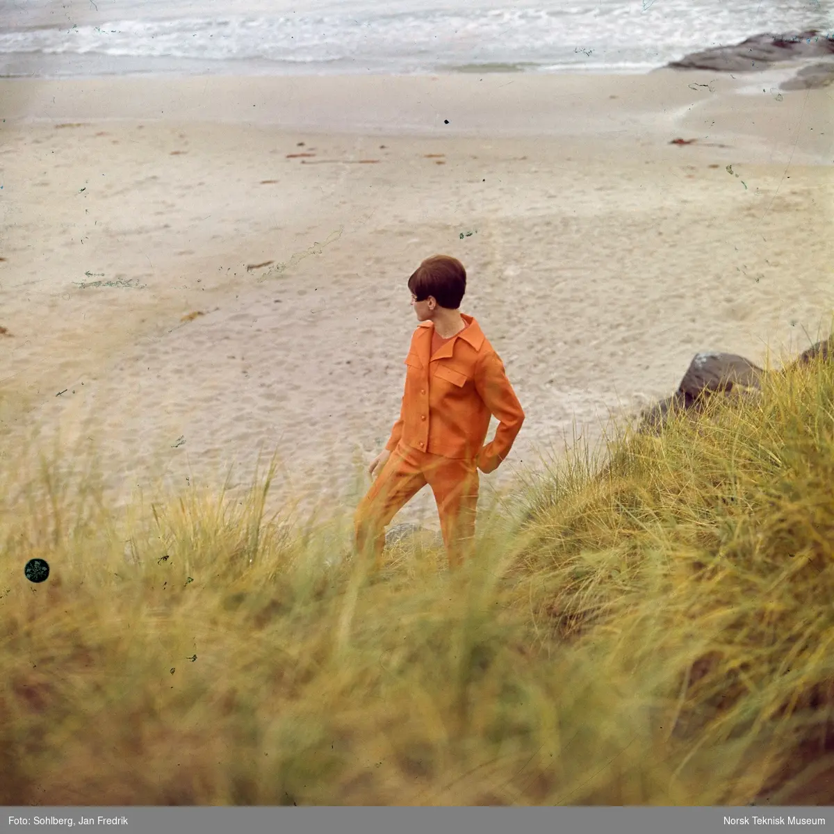 En kvinnelig modell ikledd oransje drakt med bukse og jakke er avbildet på en strand.