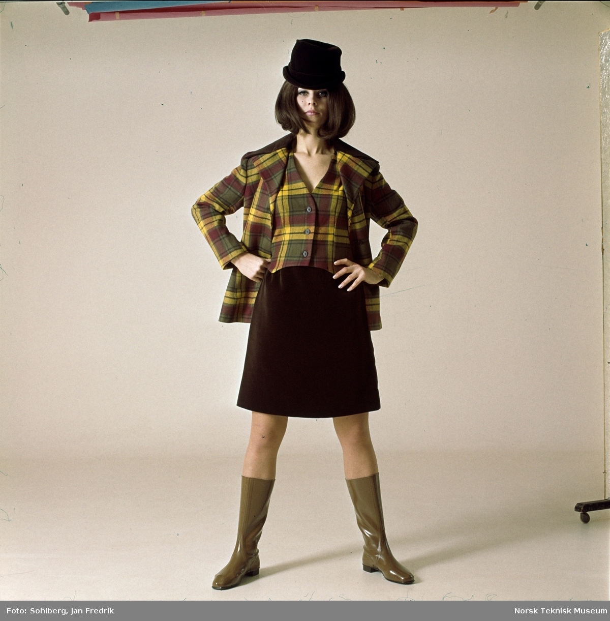 Tidlig norsk motefotografi. Kvinnelig modell med rutete jakke og vest i gult,  brunt og rødt og et mørkebrunt skjørt. Tlbehøret er en tøff hatt og lysebrune gummistøvler.