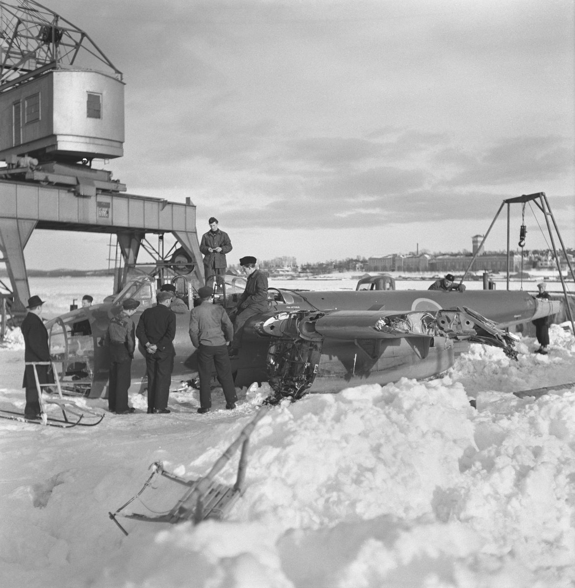 Bärgningsförsök av flygplan B 18 på isen i Härnösands hamn efter flygning i hårt väder i februari 1946. Folksamling och militär personal vid flygplanet. I bakgrunden syns lyftkran. Vy från sidan.