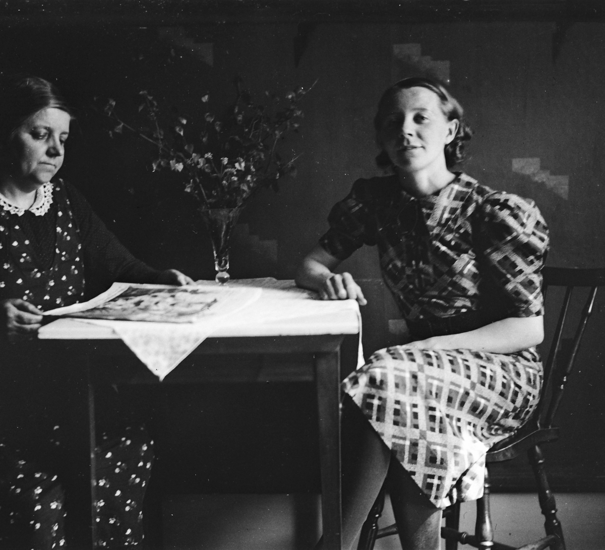 Två kvinnor sitter vid ett litet bord med duk och en vas med grönskande kvistar. Den äldre kvinnan läser en tidning. Tapeten har ett art deco-mönster.