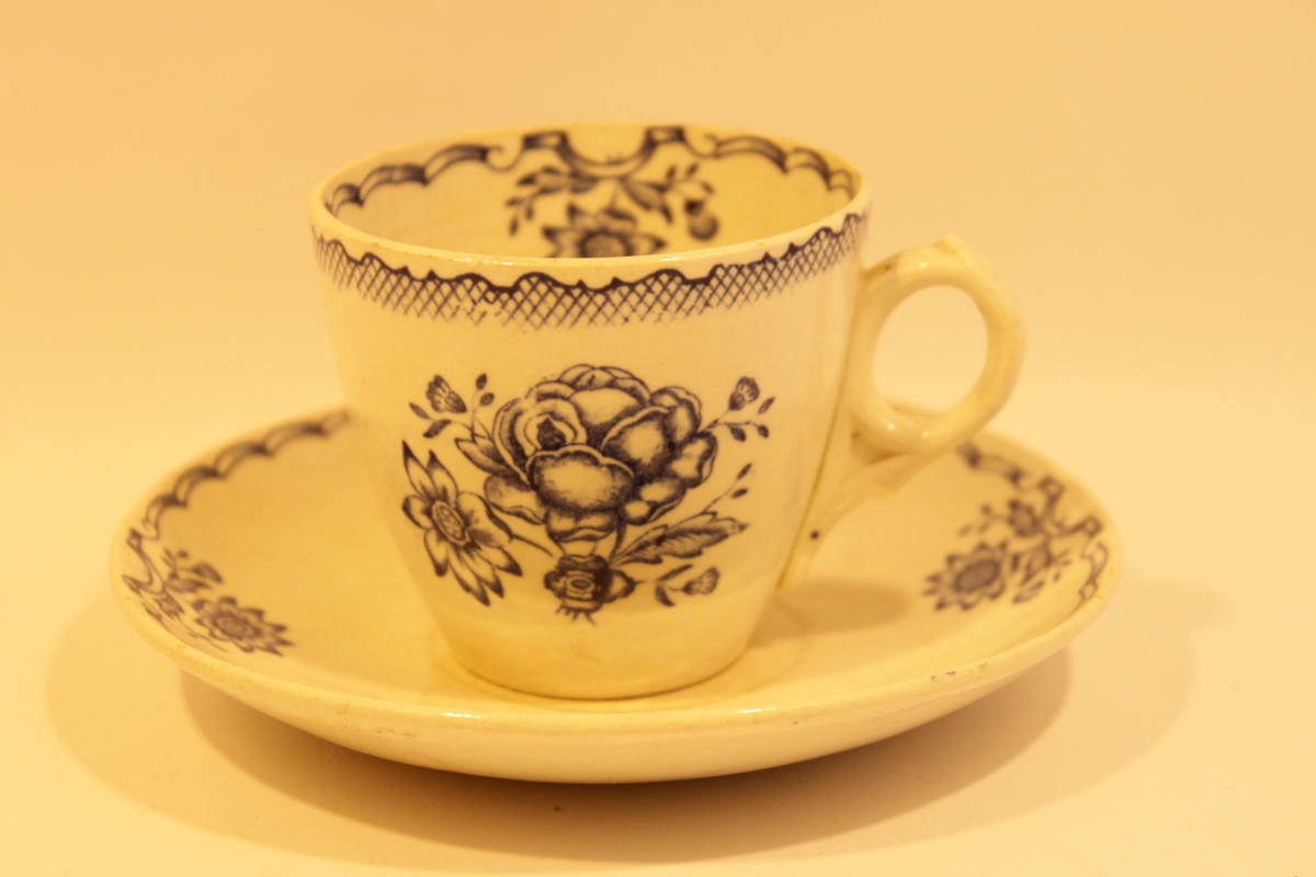 Vit kaffekopp och fat med blå dekor. Blommönster och girlander på både in- och utsidan av koppen samt på fatets ovansida. Kaffekoppen heter Bukett och tillverkades av Gustafsberg mellan åren 1936 och 1939.