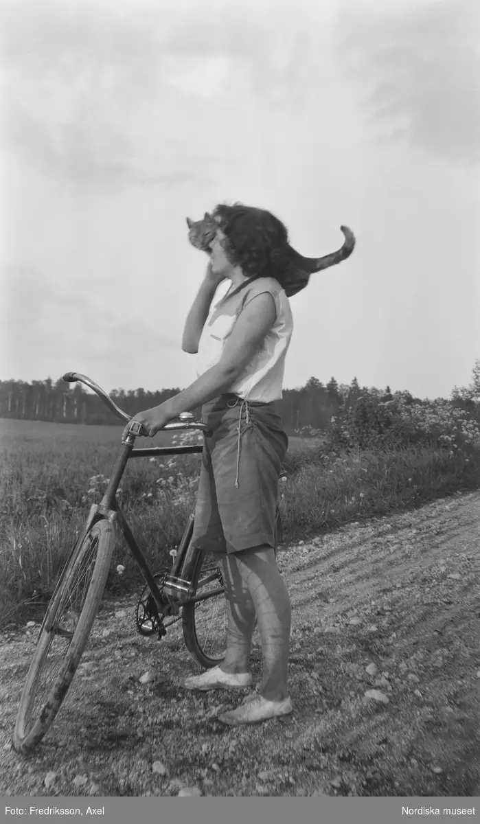 "Ladugårdselev Fogelstad med Fridas gula katt. ". En ung  kvinna i helfigur , klädd i shorts och ärmlös blus, står och håller sin cykel i vägrenen längs en grusväg. På hennes axel halvsitter en katt med svansen upp i vädret.