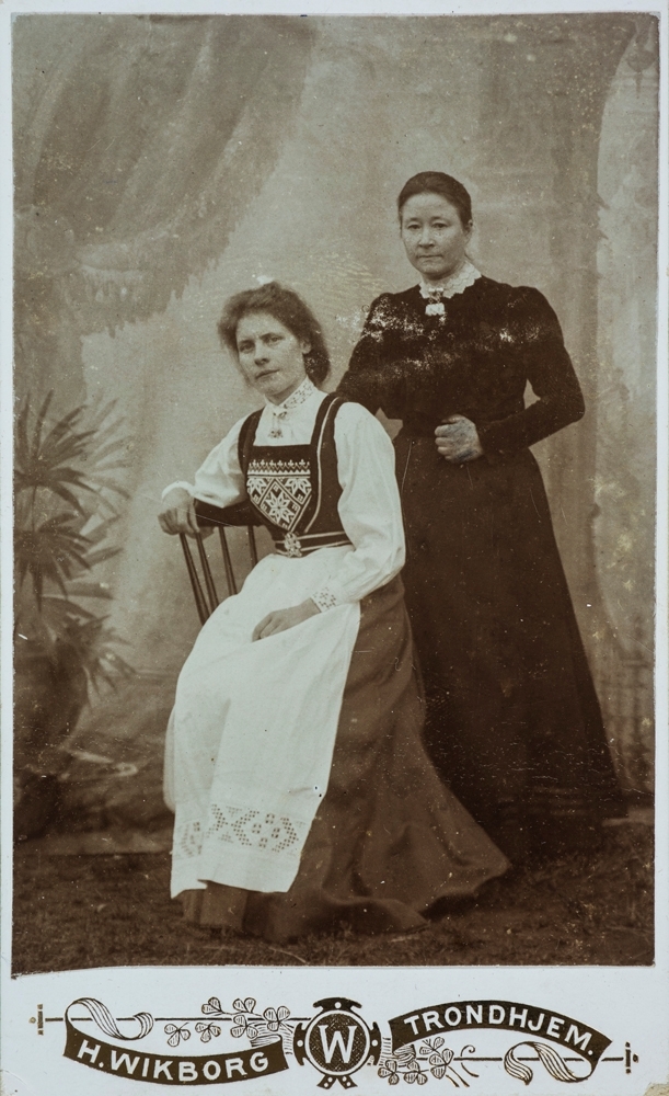 Vefsn, Mosjøen. Fotografbilde av Marta Leland og Johanna Velde. Kvinnen til venstre har nasjonaldrakt på seg (Hardangerbunad). Bildet er tatt ute med bakteppe bak.