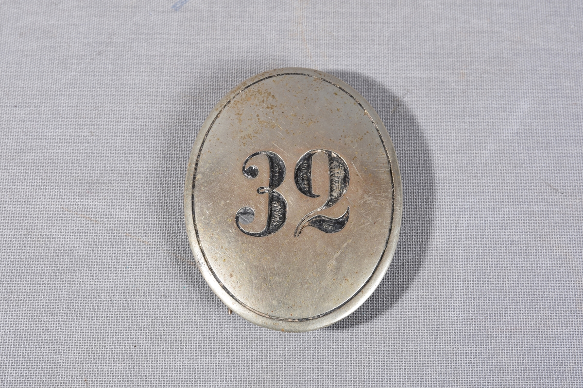 Ovalt metallskilt med konveks flate, preget nummer midt på. Branntegn for brannkonstabel nr. 32. Festes til uniform/jakke med to nåler med lås loddet på baksiden av skiltet. Stasjonsmerke i bruk fra 1863 til 1900.
