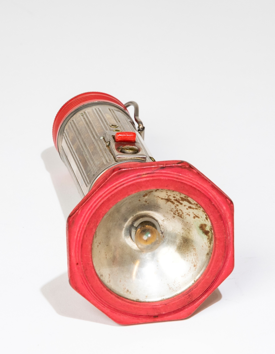 Stavlampa av plåt, gummi och glas med cylinderformad hylsa för batterier, dekorerad med räfflor. Rödlackerat skruvlock med text i relief: "SAJO". Rödlackerd, vridbar linshållare med cirkelrund glasskiva. En röd gummiring håller fast glasskivan. Hylsan är försedd med kontakter för blinkljus och stadigt ljus samt hängare av metalltråd.