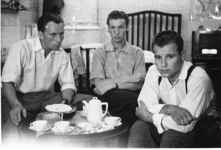 Tre amerikanska bombflygare sitter på ett rum med en kaffebricka på ett runt bord framför sig. I bakgrunden en kakelugn och en säng.