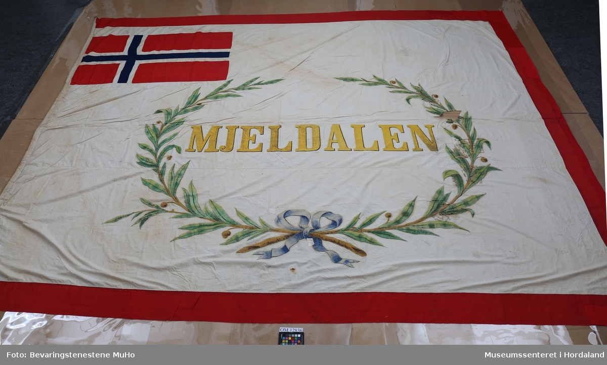 Stort flagg brukt i Mjeldalen, Osterøy. Flagget er sydd saman med handsaum av tre lengder naturfarga, tynt bomullsstoff. Det er kanta med raudt ullstoff. Flagget har påmalt dekor: Bokstavane MJELDALEN omringa av ein bladkrans med sløyfe nederst. Oppe i venstre hjørne er det norske flagget. Venstre sida er forsterka med linstoff der hampatau er tredd i til oppheng. Messingring oppe til venstre. Alt er handsauma, men med maskinsaumar der nokre nye delar raudt stoff er lappa inn.