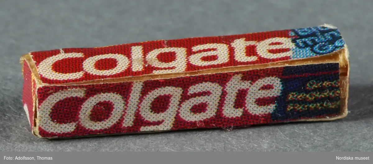 Två tandkrämstuber av märket Colgate. Den ena a) är av vitmålad avrundad metall med röd etikett och den andra b) föreställer en pappkartong och är förmodligen tillverkad av trä och papper.