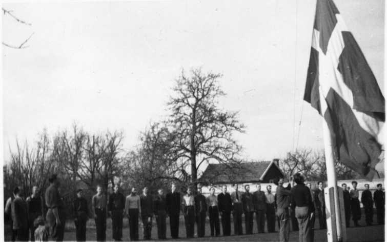 Ett led med män, unga män, stramt uppställda bakom en flaggstång där en flagga hissas/halas.