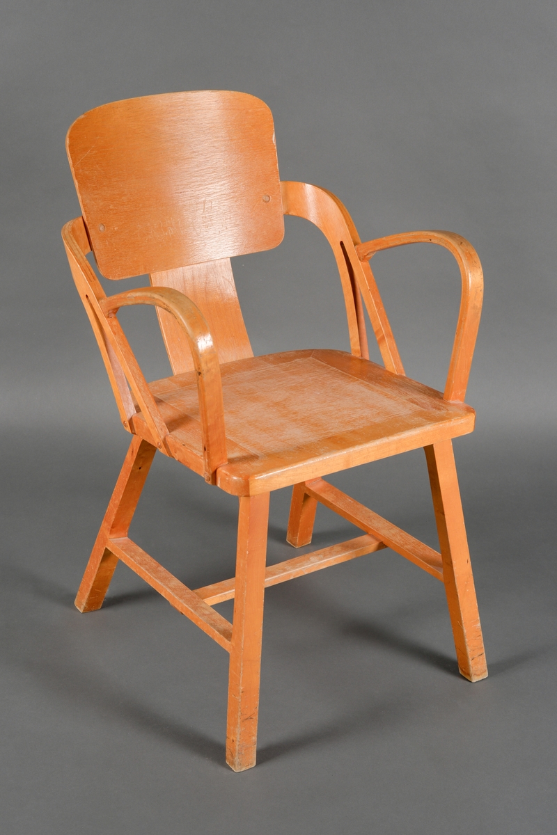 En trestol med armlener. Stolen er laget av heltre og kryssfinér. Den har sammenføyninger med lim, skruer og spiker samt treforbindelser. Setet er firkantet med avrundede hjørner. Ryggen er en firkantet plate  med avrundede hjørner. Ryggen er delvis åpen. Armelnene går i en bue fra ryggen og mot fronten. Beina er rette og firkantete. Stolen er lakkert.