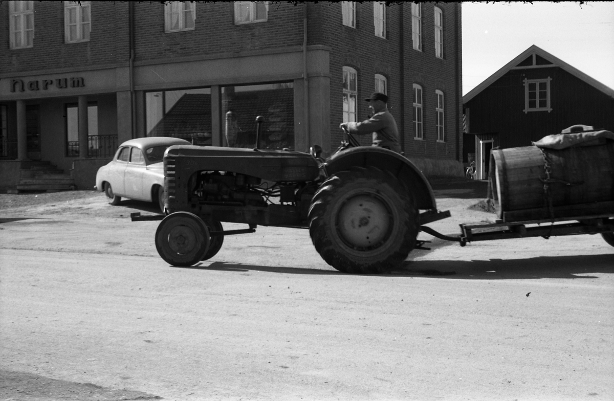 Gatebilde fra Lena april 1958. En traktor med dranktønne på tilhengeren passerer foran Narums-butikken. Mannen på traktoren er ikke identifisert.