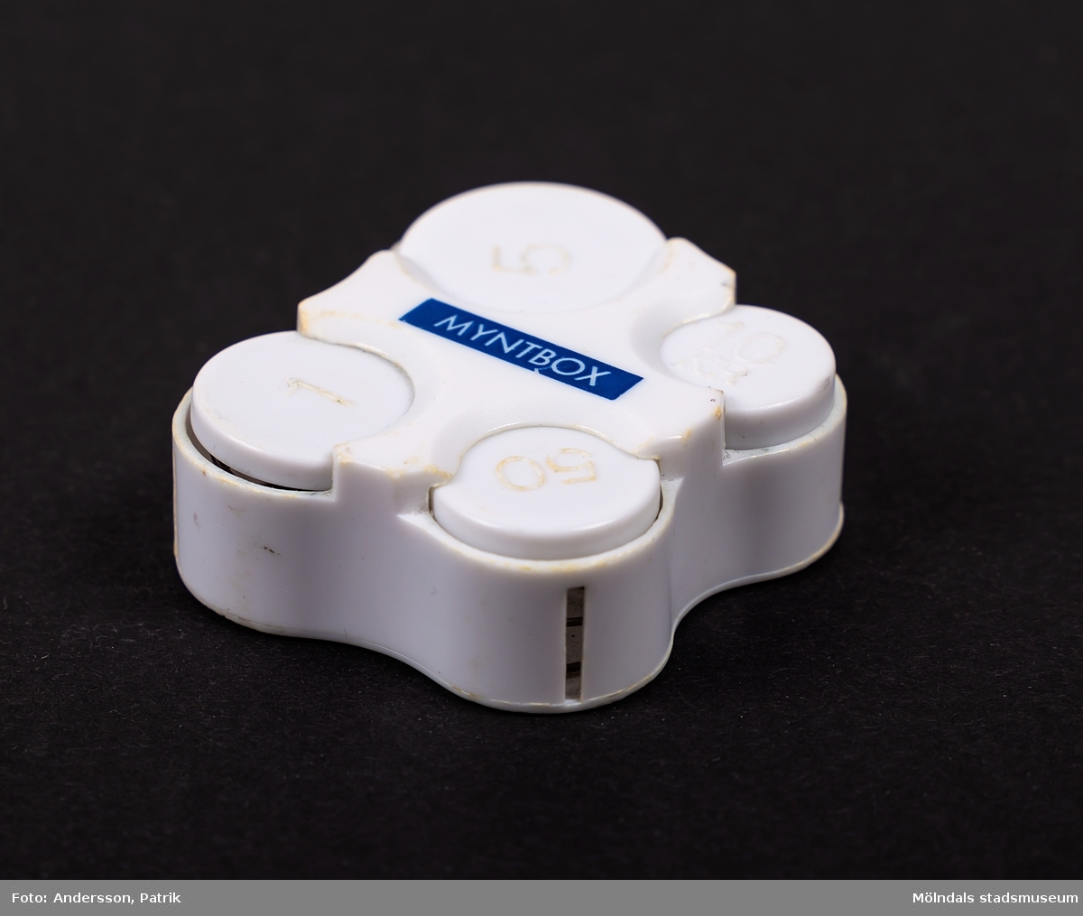 Myntbox i vit plast med texten MYNTBOX i vitt på blå botten. Mynthållaren har fack för femtioöringar, enkronor, femkronor och tiokronorsmynt. 

Tillverkad 1991 eller senare, då tiokronorsmyntet lanserades det året.