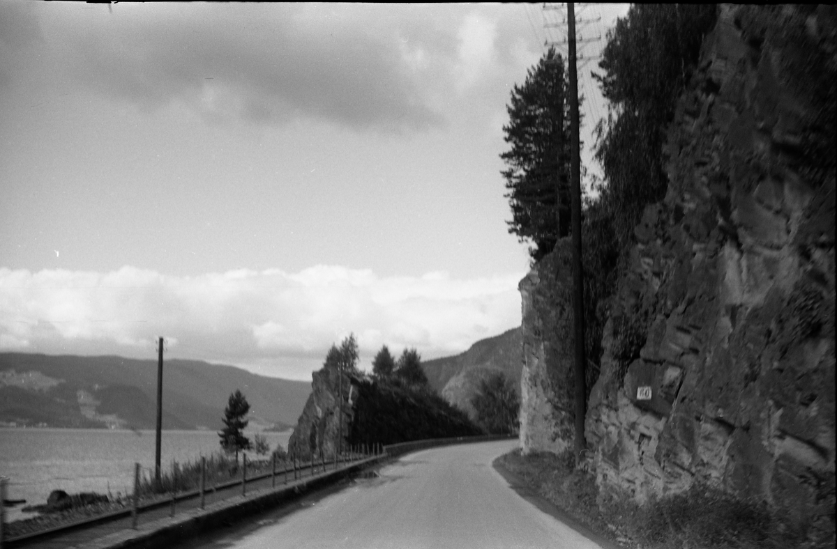 Veg gjennom fjellskjæring. På nedsida av vegen renner ei elv. Trolig mellom Leira og Fagernes 1958. Strandefjorden og skinnegangen til Valdresbanen til venstre i bildet.