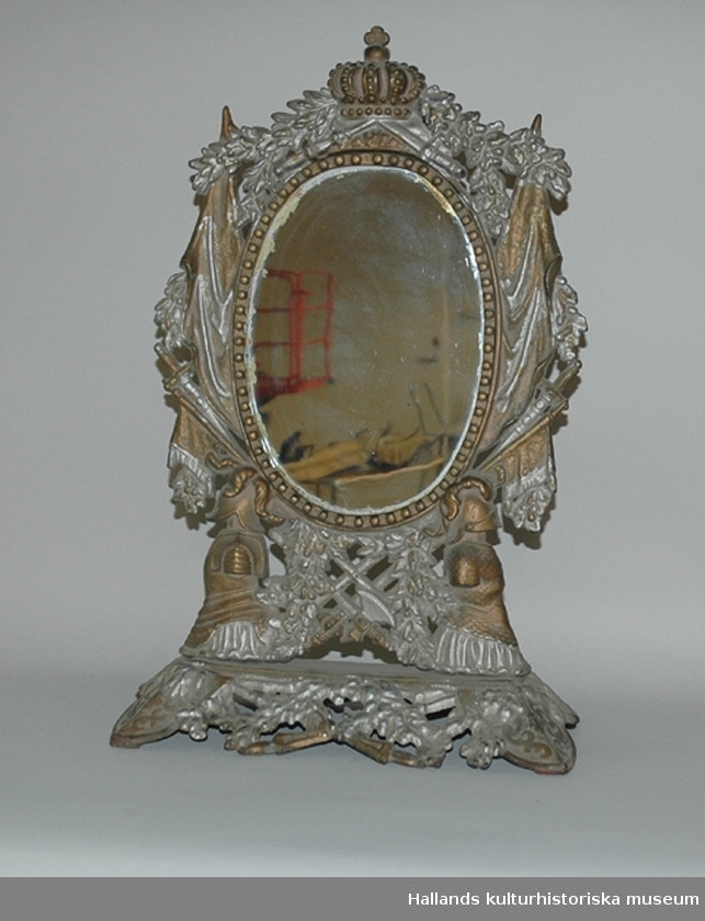 Oval spegel. I ställ av gjutjärn, målat i brons och silver. Total höjd 51 cm, total bredd 33,5 cm, glasets höjd 24,8 cm, glasets bredd 17,1 cm.