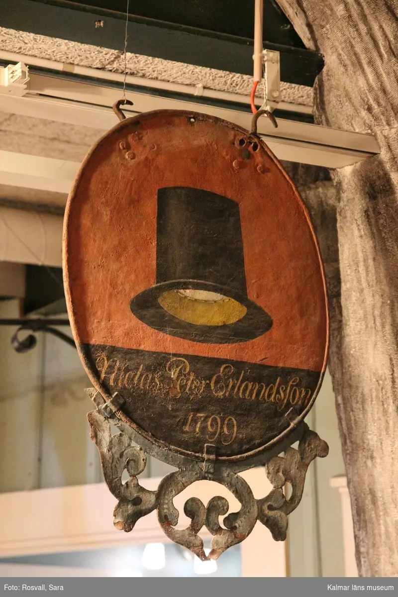 KLM 5658. Skylt, hattmakarskylt. Oval skylt av järnplåt, bemålad, på ena sidan en svart hatt på röd botten, på den andra en svart hatt på blå botten. Nederst på båda sidor med gula bokstäver på svart botten:  Niclas Peter Ehrlandsson 1799 .