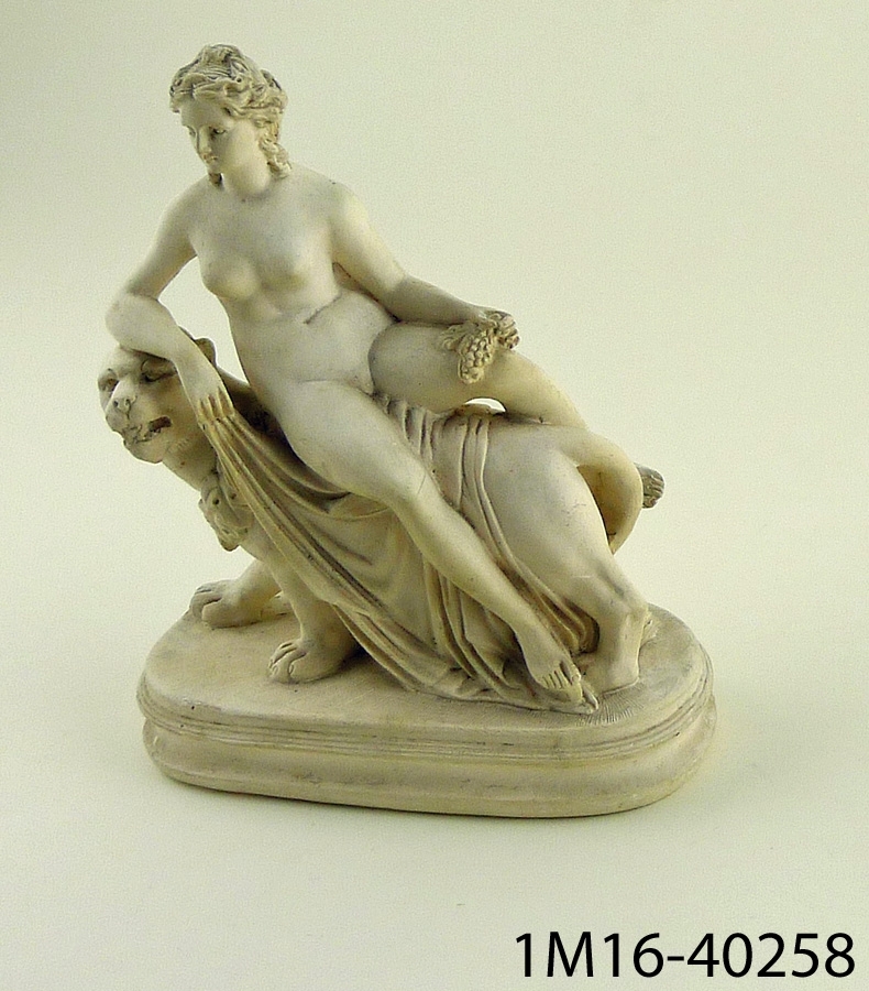 Gipsskulptur föreställande naken kvinna sittande på lejoninna. Kvinnan sitter på ett tygstycke och håller en vindruvsklase i sin ena hand.