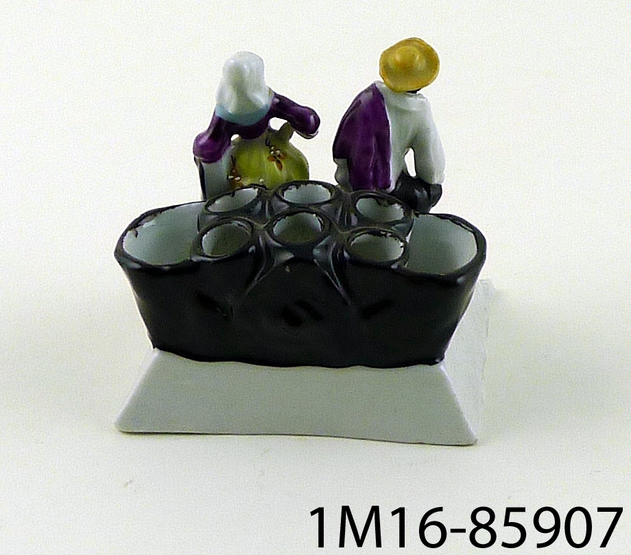 Figurin föreställande en kvinna och en man stående framför tre stycken behållare för blommor.