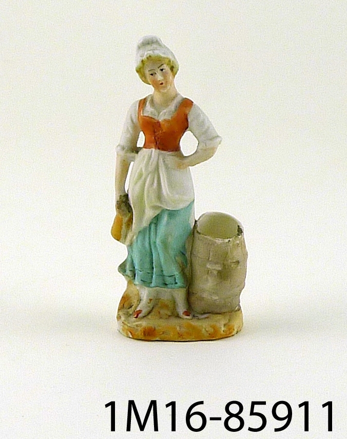 Figurin föreställande kvinna i vitt med hatt och rött livstycke, vit blus och vit uppskörtad kjol med grön underkjol, hållande ett krus i handen stående vid en ihålig tunna. Tunnan kan användas som vas.