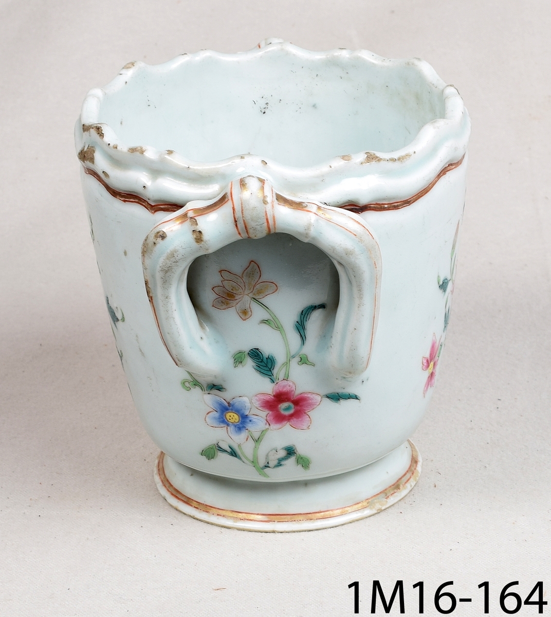 Rund kruka av keramik med ljusblå glasyr och handmålad blomdekor i många färger och guld. Krukan har två hänklar i överkant och en vågformad kant.