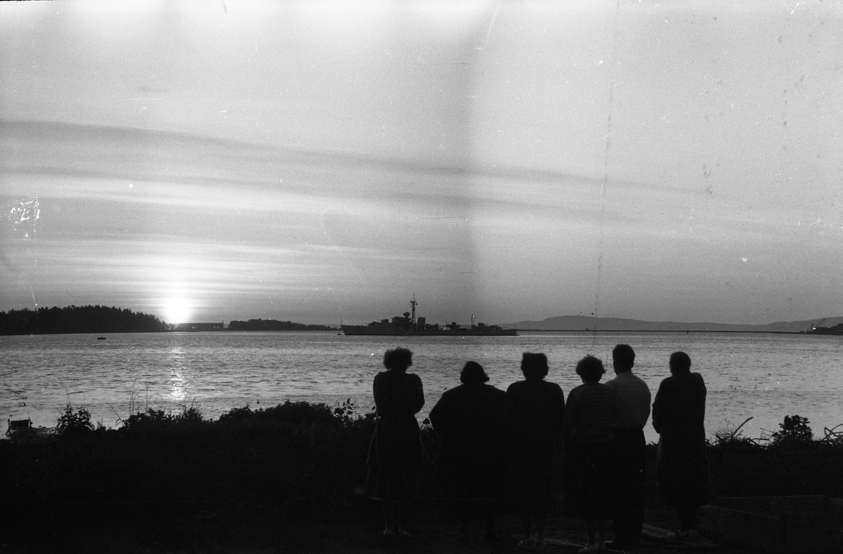 To avfotograferte bilder av ei gruppe personer samlet ved en fjord i solnedgang eller muligens midnattsol. Et fartøy passerer ute på fjorden.