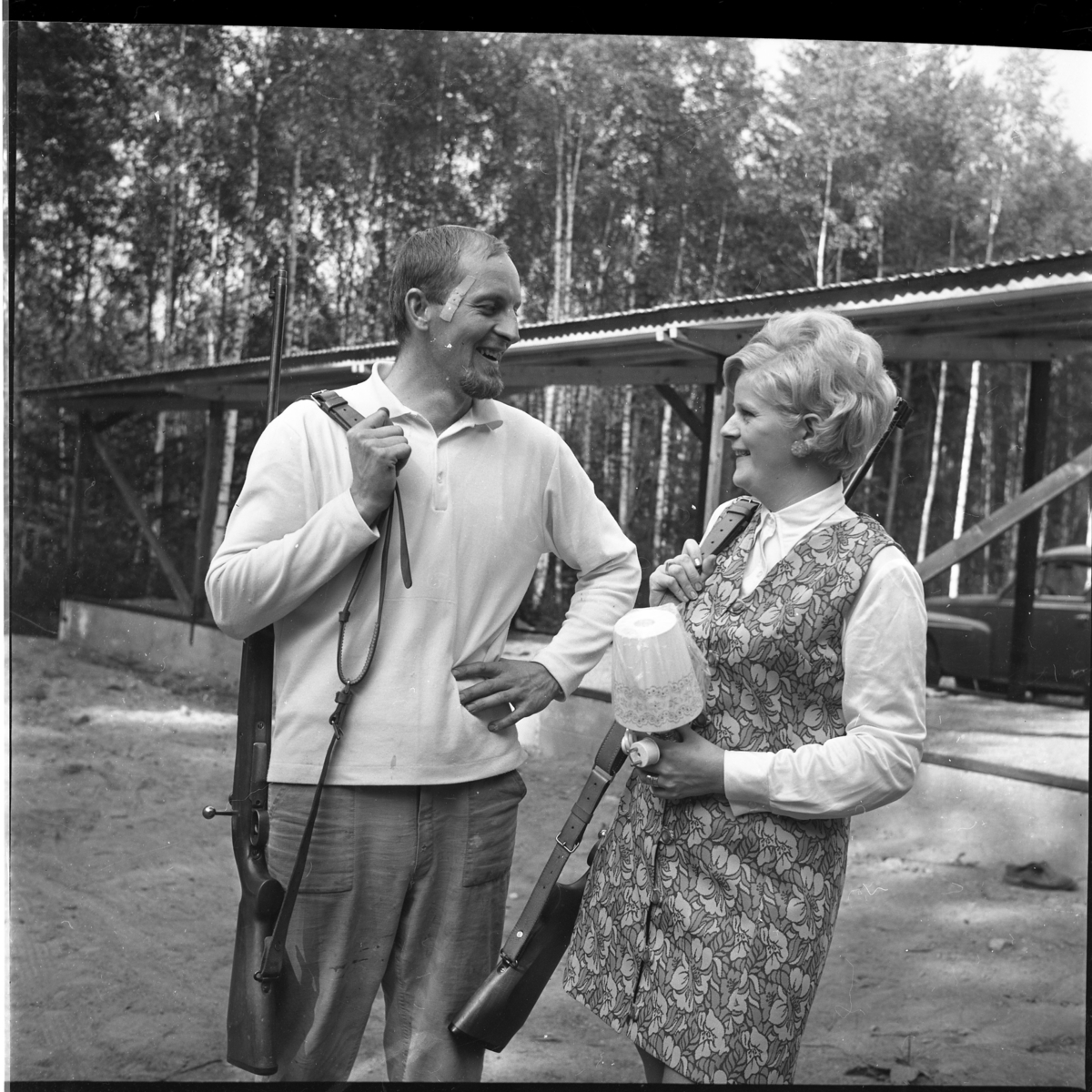 En kvinna och en man står vid en skjutbanan med gevär i rem över axeln. Det är Åke Gustavsson och Inga Lena Karlsson. Hon håller en bordslampa i handen - troligen ett pris i skyttetävling. De är vända mot varandra och skrattar.