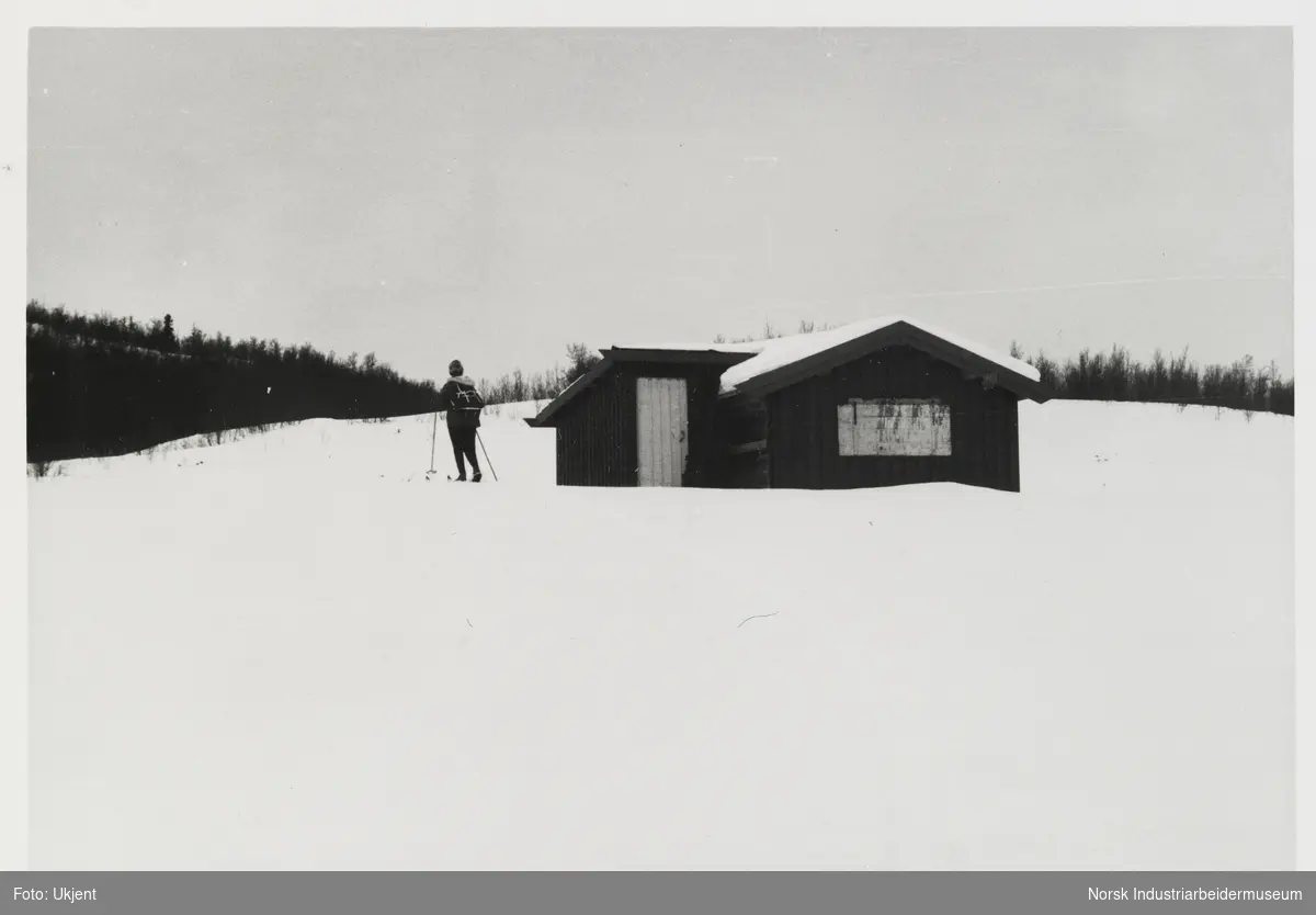 Grønli. Person med ryggsekk og ski på beina står ved siden av liten hytte i snødekt landskap