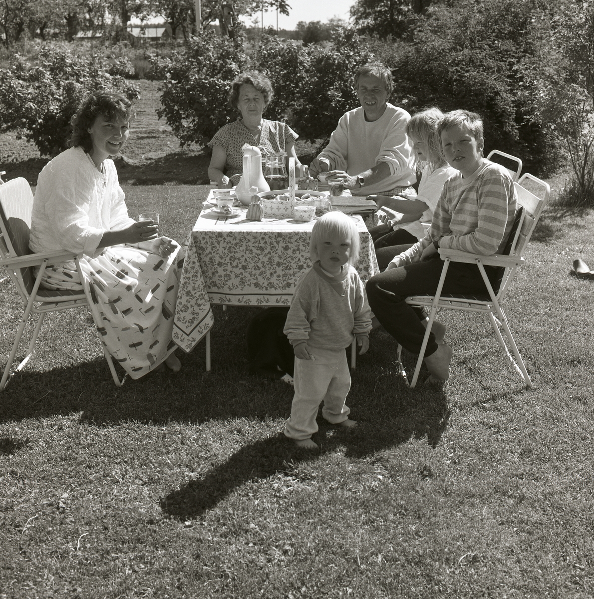 Ute i trädgården sitter Adéle, en man, en kvinna och barn vid ett fikabord, på bordet ligger en duk. Det finns även stolar, buskar, gräs och träd. Från Adéles 70-årsdag 16 juni 1988.