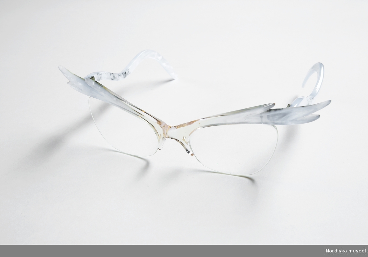 Ett par glasögon med pärblå "vingad" kant, troligen av plexiglas. Nylontråd fungerar som en kant under glasen. Trådarna är fästa vid näskuddarna samt på frontens baksida, resten av trådarna (ungefär 7 cm) hänger fritt på baksidan. Oslipade glas med sadelbrygga. Vågformade skalmar i samma pärlblå färg. 
/Petrine Knight, 2015-07-01