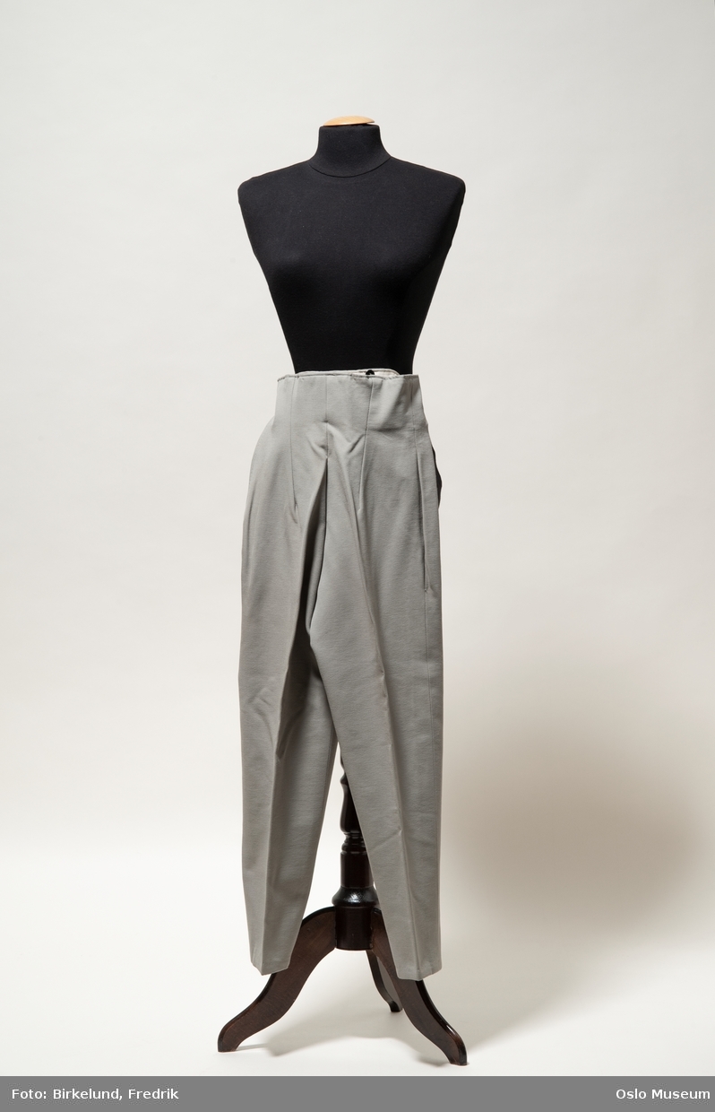 Lange grå bukser i elastisk, syntetisk stoff. Lukkes med glidelås i siden.