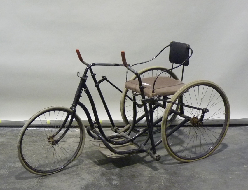 Rullstol av svartmålade järnrör på cykelhjulskonstruktion. Stoppad sits. Styranordning för framhjulet. Drives med handkraft: spakar vid sidorna.