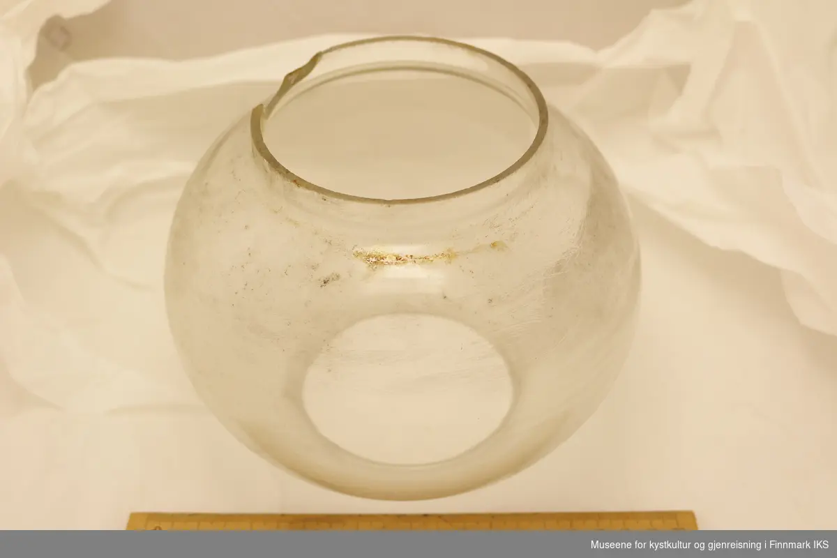 Lampeskjerm i glass med rund form. Åpen i topp og bunn, og har sylinderformet kant langs åpningene. Lampeskjermen er skitten, og har hakk langs kanten i den ene enden.