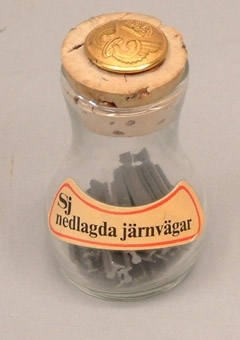 Liten glasflaska innehållande små bitar av skalaräls.
Gul etikett med texten "SJ nedlagda järnvägar". På korken är fastsatt en uniformsknapp från 1973 års reglemente.