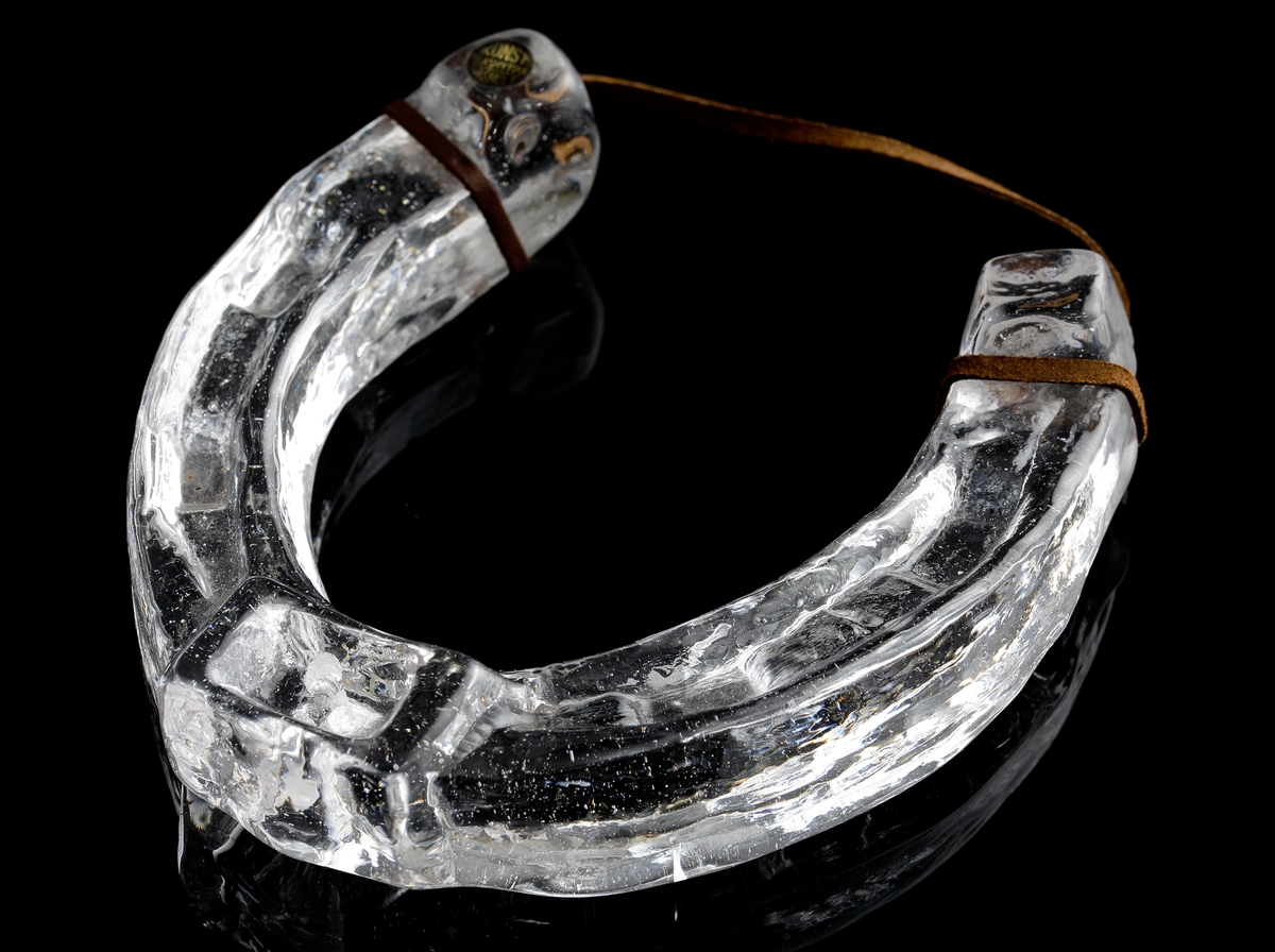 Hästsko i ofärgat glas med läderrem för montering på vägg.
Etikett: Rund svart med gul text "Konstglashyttan Urshult".