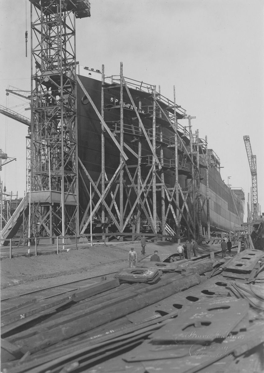 Motivet viser MT SPINANGER (bygget i 1927) under bygging ved Schiechau i Gdansk. 6 store havnekraner rundt skroget som ligger i tørrdokk.