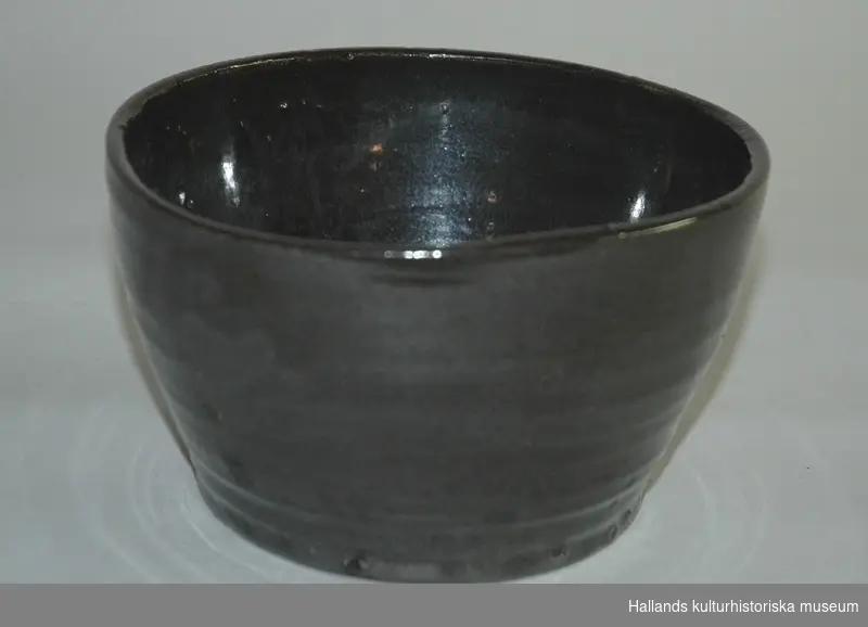 Skålen är av bränd keramik. Brun-svart färg, glaserad. Märkt i botten "Cederholm, Varberg."