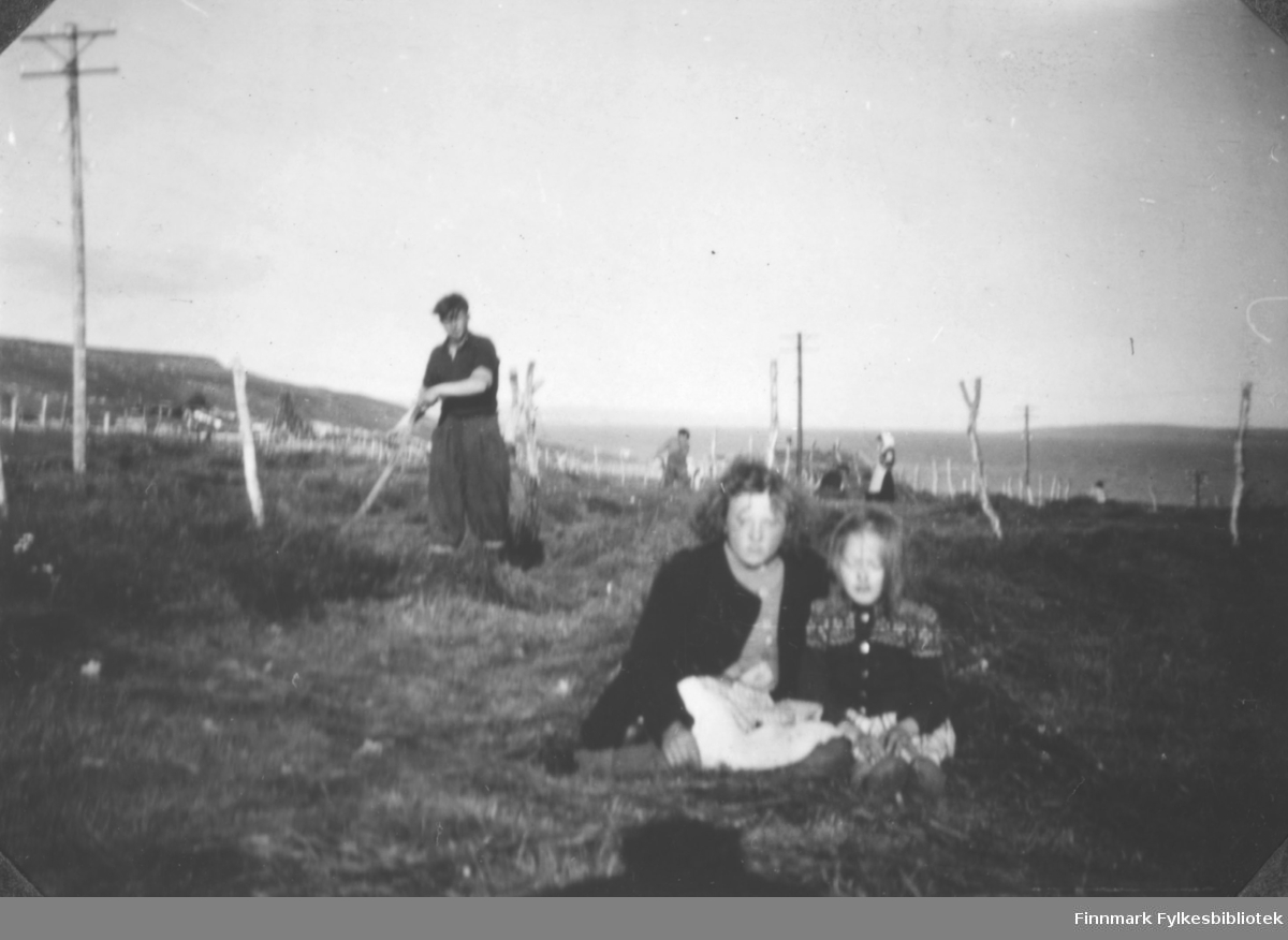 Flere personer fotografert på et jorde i Karlebotn i Nesseby. Sigrid Margit Mathisen (til venstre) og Anna Agnethe Matisen sitter på bakken foran i bildet. Mannen i bakgrunnen er ukjent.