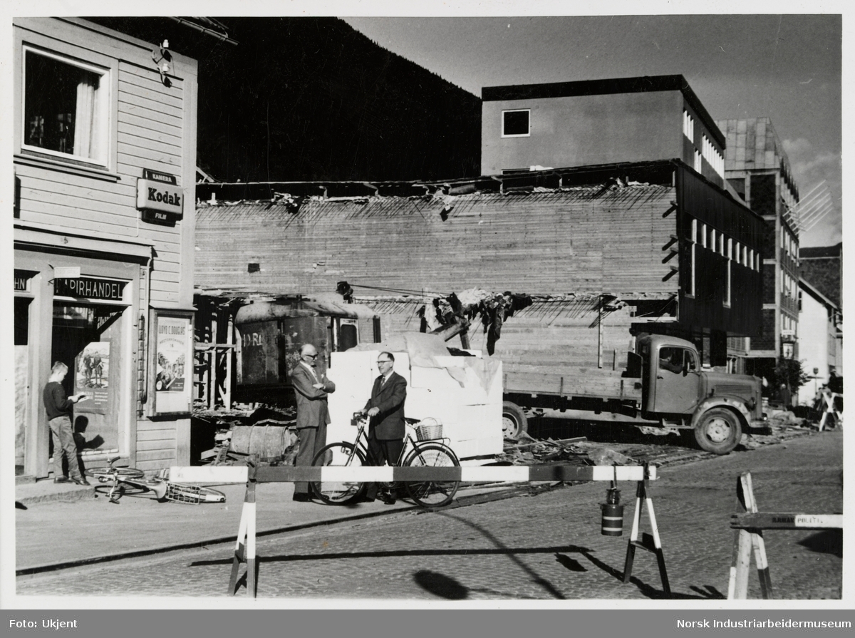 Otto Aubert og James Coward på sykkel ved varehuset, 2. byggetrinn. Gutt står ved døråpning til papirhandel og leser bok, sykkel ligger på bakken. På husfasade er skilt for Kodak kamera og film.