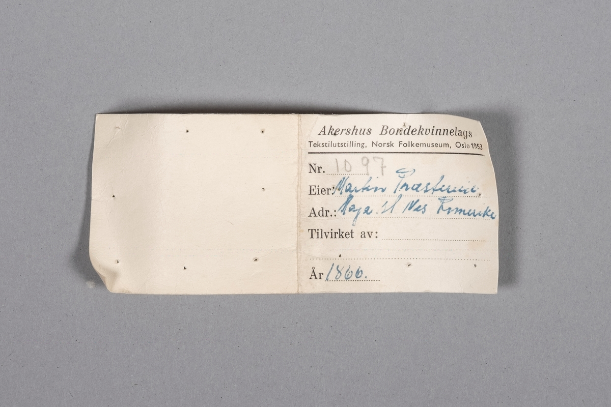 Etikett som ble brukt til tekstilutstillingen på Norsk Folkemuseum i 1953. Rektangulær, brettet i to, med skrift på fremsiden. Opplysninger om hvem som eier dåpskjolen, hvor eier bor, hvem som har produsert den, og når den er produsert.
