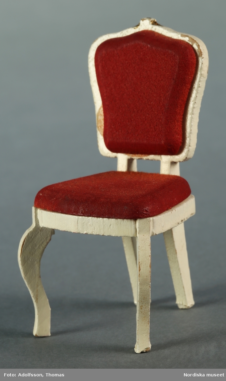 Ett par vita stolar av trä med röd stoppning, tillverkade av Lundby. Efterliknar stilmöbler i nyrokoko. Stolarna är placerade i matsalen vid väggen mot serveringsgången, på ömse sidor om ett vitt matbord (NM.333314) i dockskåpet NM.331721+.
