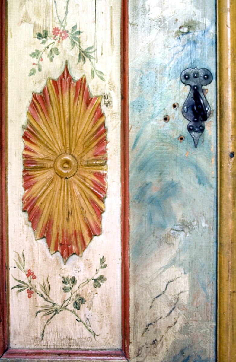 Skåp av furu. Bred profilerad rak krönlist. Två dörrar, med låset placerat på vänster dörrhalva. Framsidan är rikt dekorerad med bland annat utskurna och målade dekorelement av trä målade i gulbrunt och rött. På dörrarna listverk målade i gulbrunt. På förhöjningar utanför listverket pålagda dekorelement i form av eldslågor målade i rött. På dörrspeglarna pålagda dekorelement med utgående strålar målade i gulbrunt och rött, omgivna av målade korslagda kvistar. I övrigt är skåpet marmorerat i blå och vita toner. Nedre delen av skåpet målat i rosa och gulbrunt och partiet mellan frambenen svängt. Invändigt tre hyllor, varav en skedhylla. På den mellersta hyllan hänger en draglåda. 
Invändigt omålat liksom höger utsida, då skåpet är tänkt att placeras i ett hörn.
