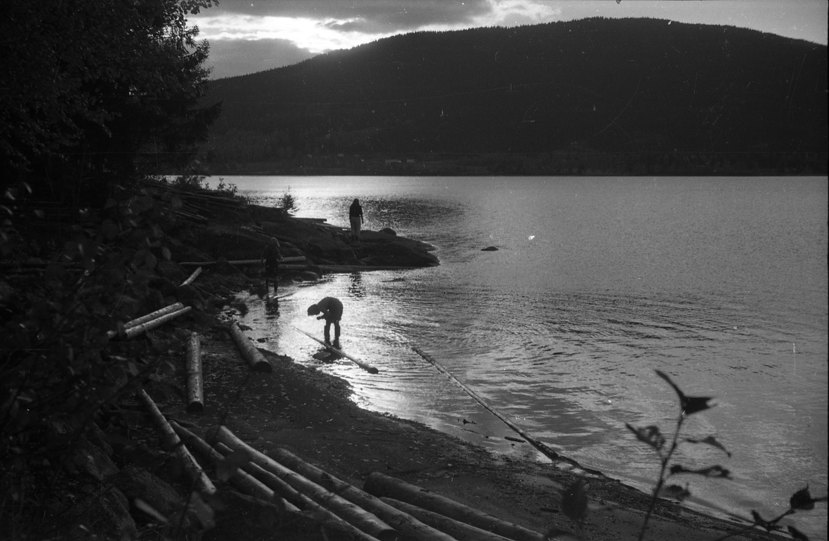 Avfotografert bilde fra stranda av en innsjø. Et par personer i eller ved vannet. Stedet er ikke identifisert.