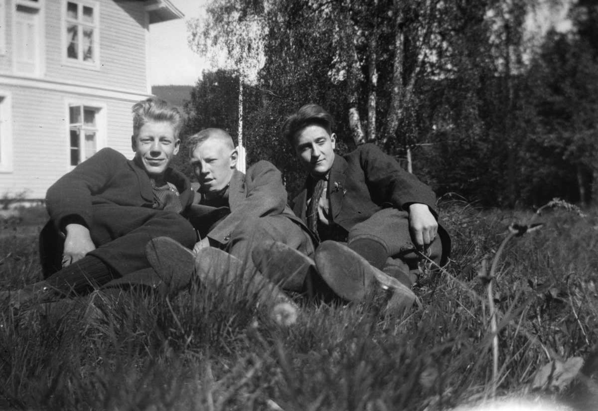 Tre unge menn i gresset, med et hus i bakgrunnen.
